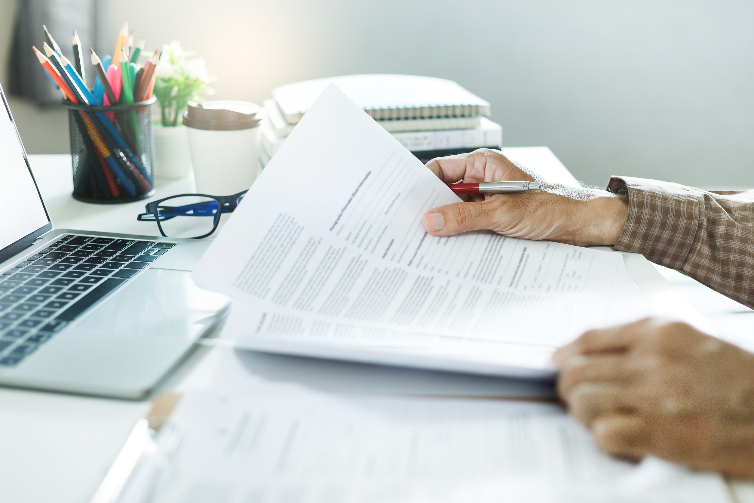 Seated at a desk, a person reviews insurance policies with a pen in hand. Nearby are a laptop, eyeglasses, a plant, and a cup of colorful pens. Sunlight filters in, creating a bright and organized workspace perfect for considering how often to assess these important documents.