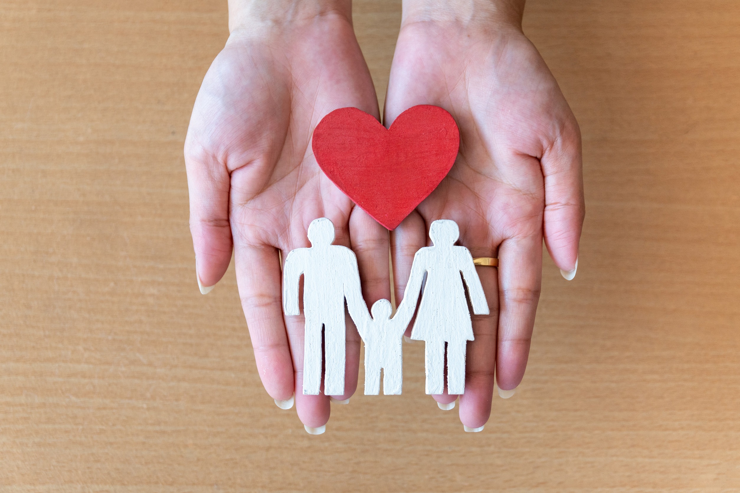 Two hands holding a red heart and white wooden family cutouts, symbolizing love, care, and the protection that life insurance can provide on a wooden surface background.
