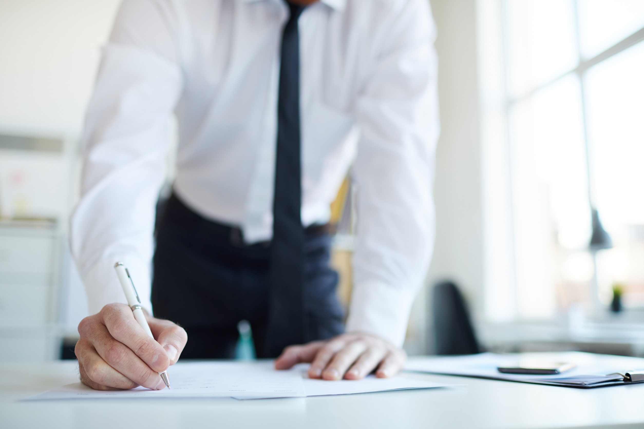 A person in a white dress shirt and dark tie leans over a desk, writing on a document with a pen—reflecting the dedication of independent insurance brokers to delivering superior value in a bright office filled with papers and a smartphone.