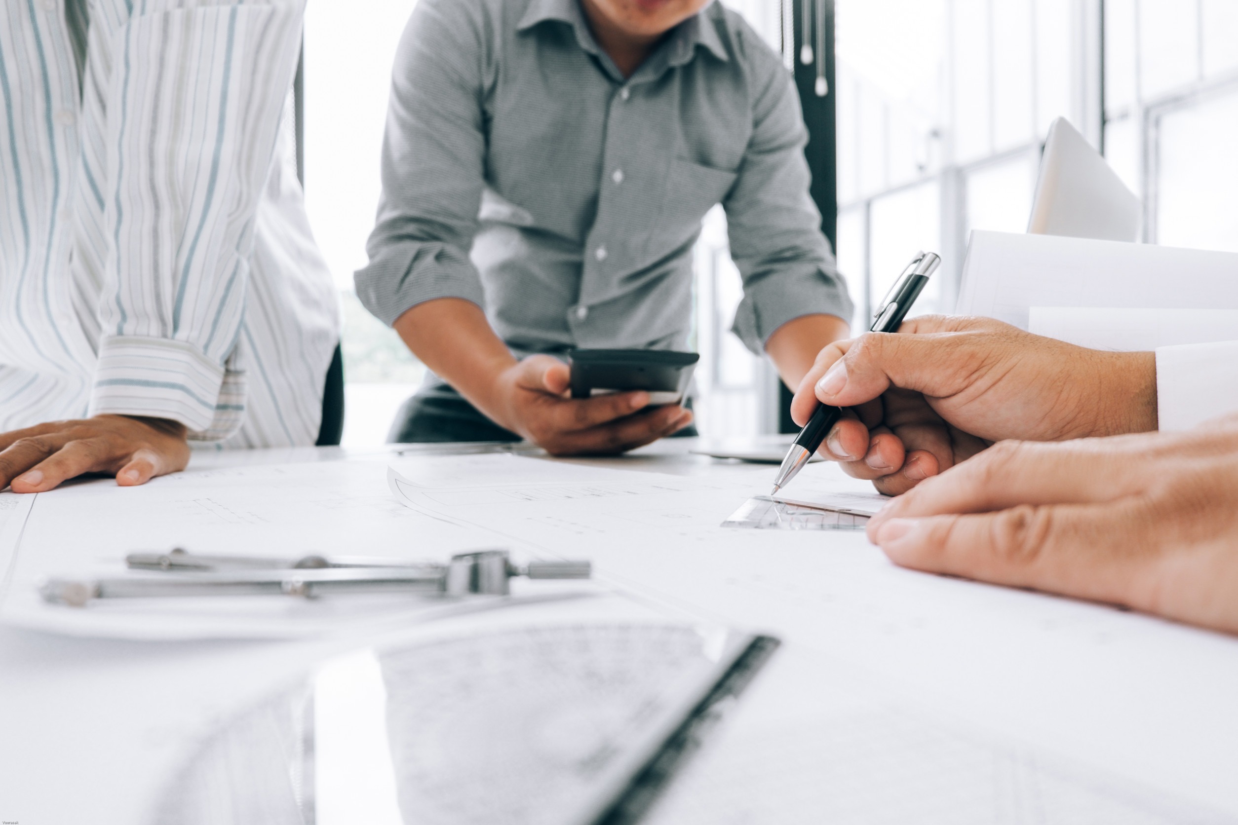 Three people in business attire collaborate at a table covered with documents, a ruler, and a compass. One writes notes for National Insurance Brokers, another uses a phone for Hice Agency, and a third stands nearby leaning over the table.