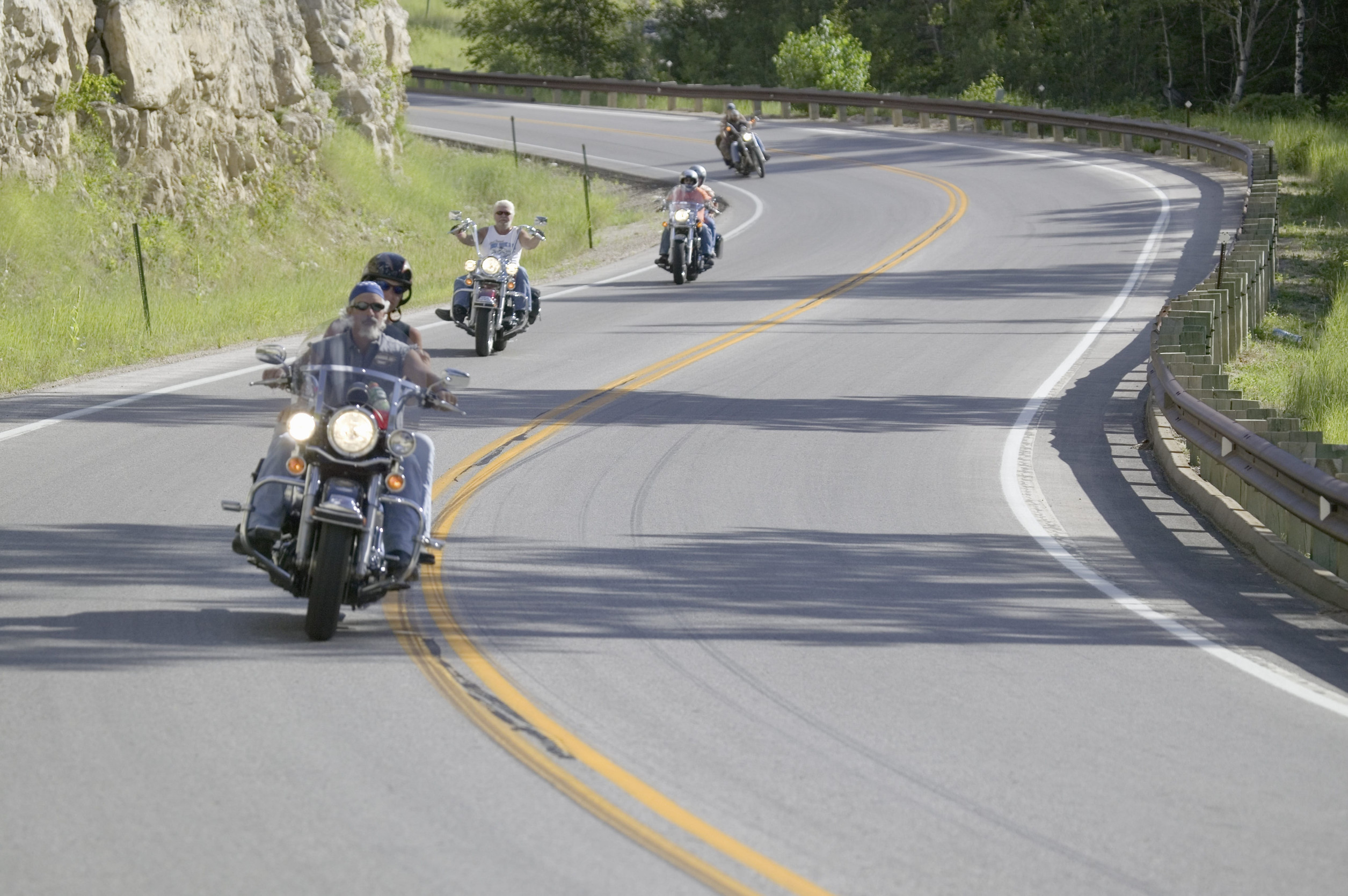 Four motorcyclists ride along a winding, two-lane road bordered by trees and rocky cliffs on a sunny day. The group is spread out along the curve, showcasing how to ride safely while enjoying peace of mind with recreational vehicle insurance.