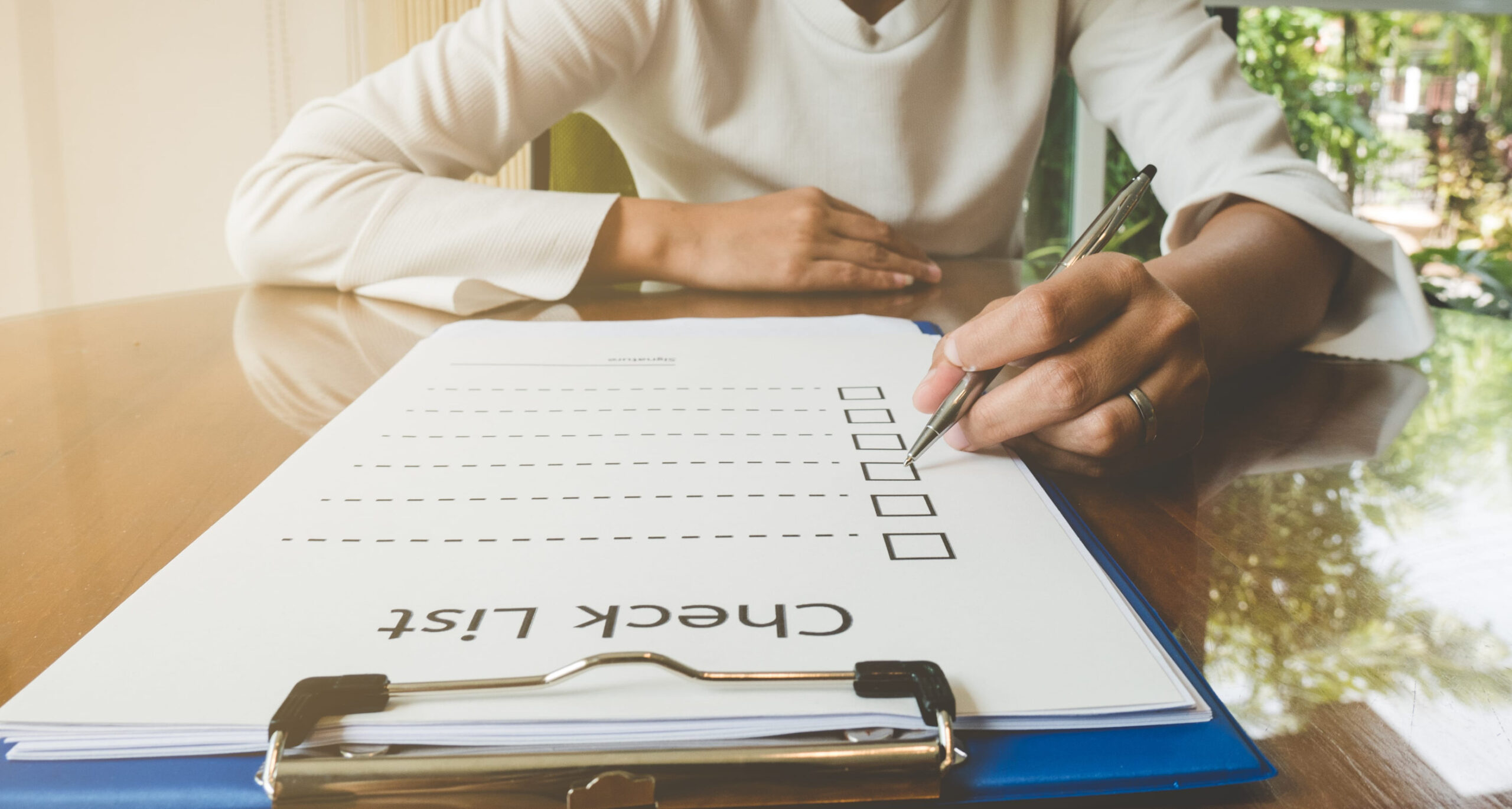 A person sits at a table, filling out a Fall Insurance Checklist on a clipboard with a pen. The checklist has empty boxes and the words “Check List” are visible. Sunlight comes through a window in the background, suggesting it’s time for a fall insurance review—especially important for Minnesota insurance policyholders.
